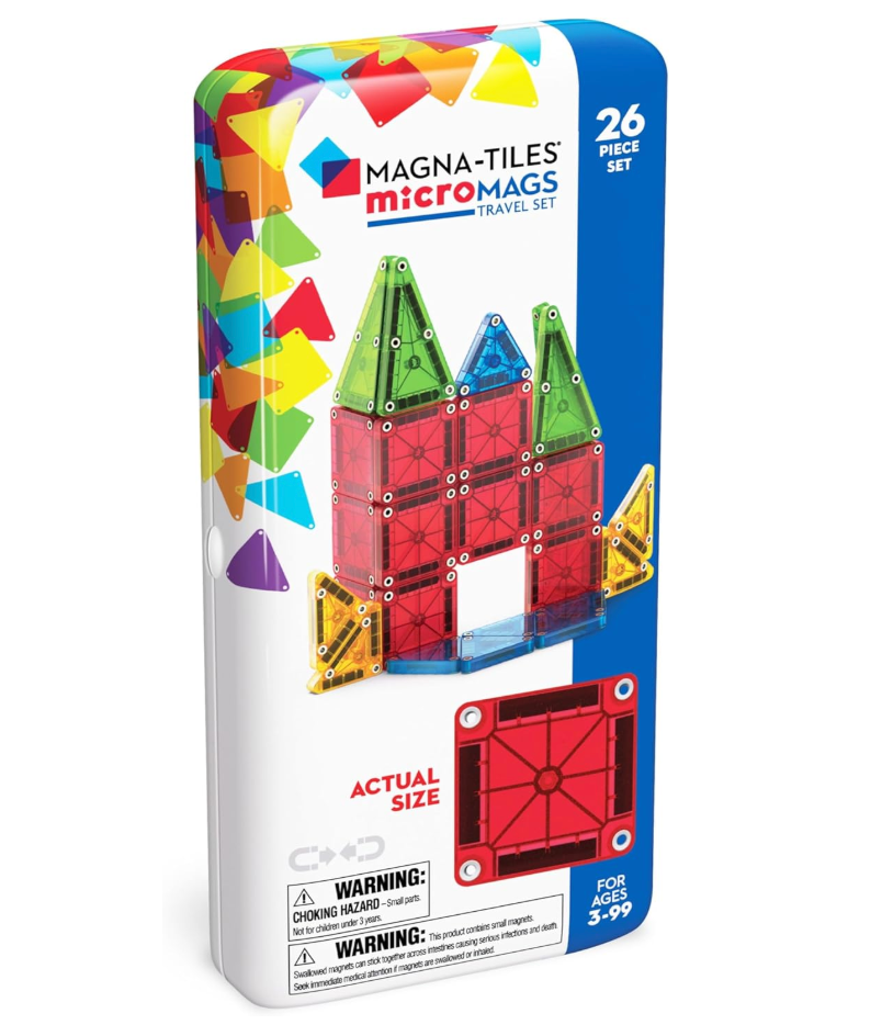 Three Magna-Tiles sets arranged on a wooden surface — Classic 32-Piece, Stardust 15-Piece, and microMAGS Travel Set — with colorful translucent and iridescent tiles scattered around them in soft natural light.