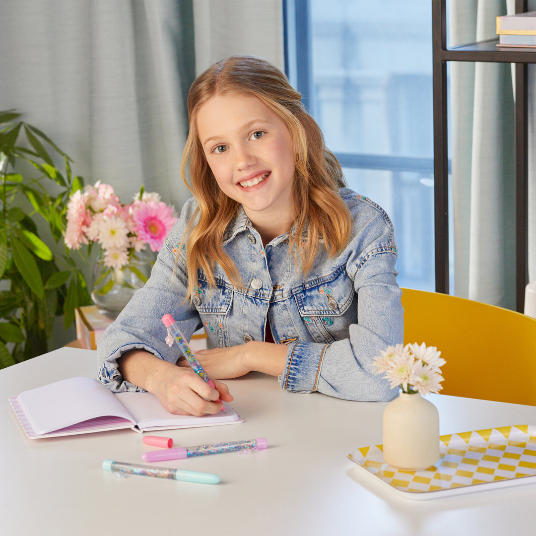 Young girl writing in a notebook at a table with flowers and stationery.