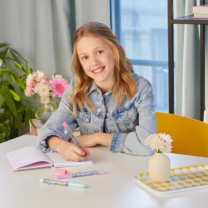 Young girl writing in a notebook at a table with flowers and stationery.