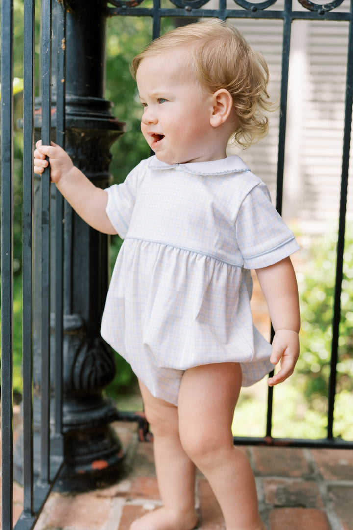 Child in a light blue and yellow checked bubble outfit standing outdoors near a black metal gate.