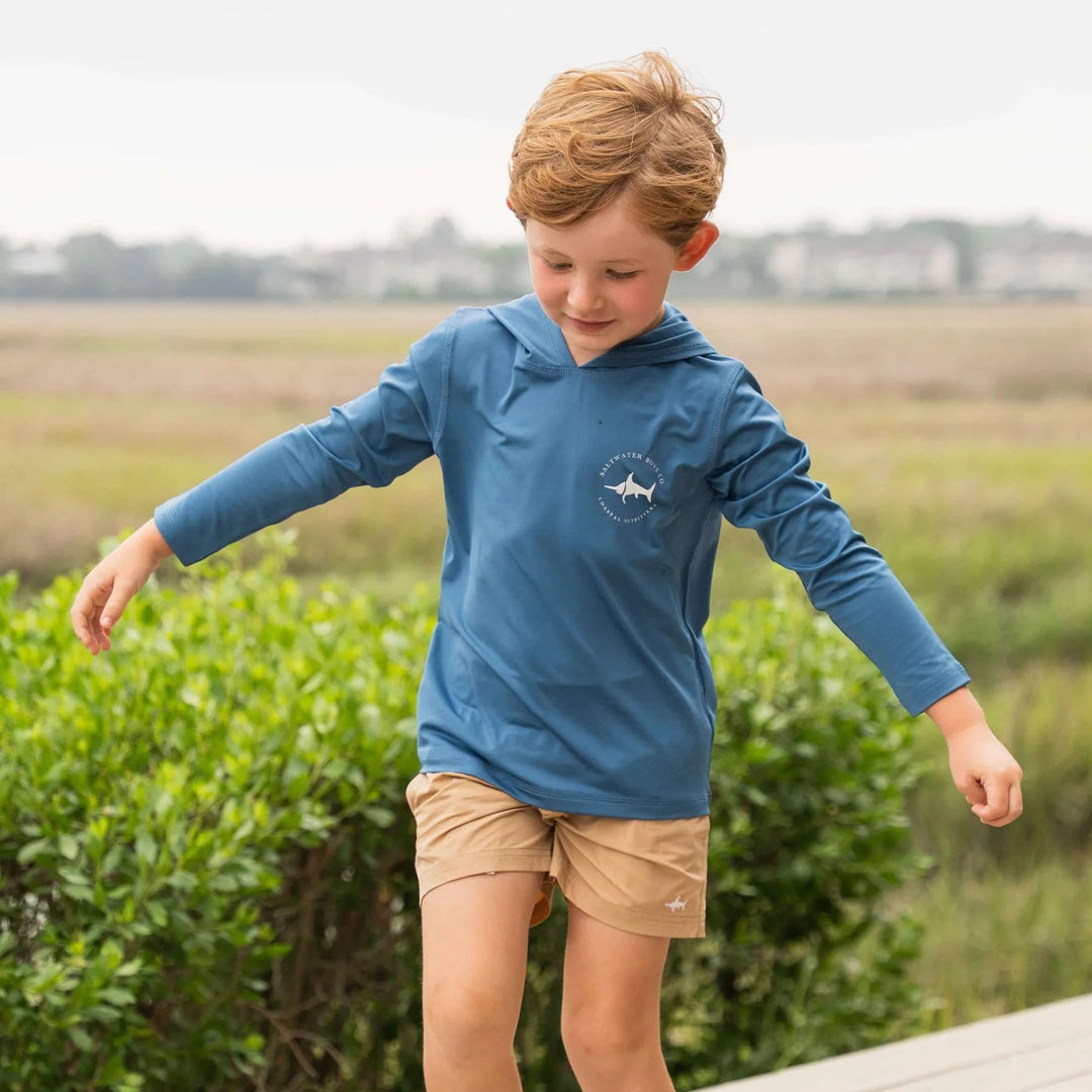 Child wearing a blue shirt and beige shorts standing on a wooden platform with greenery in the background